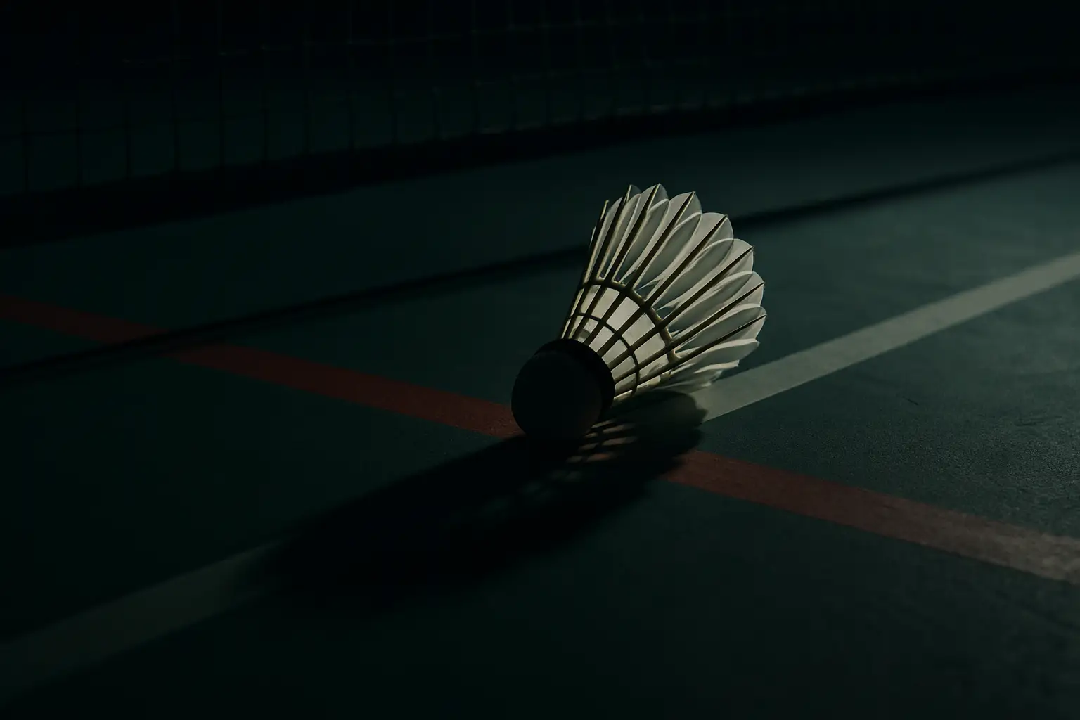 A badminton shuttlecock on a court floor near a net with a blurred background suggesting tension